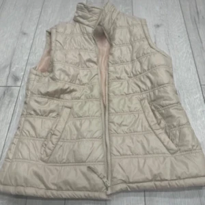 Beige quiltad dunväst med dragkedja - Snygg beige dunväst med quiltad design och hög krage. Västen har två fickor framtill och stängs med dragkedja. Perfekt att slänga över en hoodie eller tröja för extra värme under kyliga dagar.