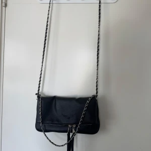 Svart axelväska med kedjerem - Zadig inspirerad. Snygg svart axelväska i skinn med silverfärgad kedjerem och dragkedja framtill. Väskan har ett rymligt huvudfack och innerfoder i brunt tyg. Perfekt storlek för det viktigaste och ger en cool vibe till din outfit.
