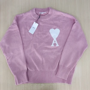 Rosa stickad tröja från Ami Paris - Säljer en ljusrosa stickad tröja från Ami Paris med rund hals och ribbade muddar. På bröstet finns ett vitt hjärta med bokstaven A. Tröjan har lång ärm och är perfekt för dig som gillar snygga detaljer och mjuka material.