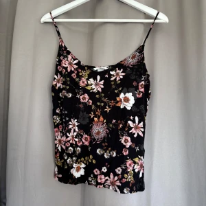 Blommig svart linnetopp från H&M - Svart linnetopp från H&M med tunna axelband och ett färgstarkt blommigt mönster i rosa, vitt och grönt. Toppen har en rak passform och ärmlös design, perfekt för att ge din outfit en cool och fräsch vibe. Materialet är mjukt och lätt, vilket gör den skön att bära.