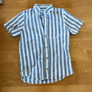 Randig kortärmad skjorta Jack & Jones - Snygg kortärmad skjorta från Jack & Jones med breda blå och vita ränder. Skjortan har klassisk krage med knappar och är tillverkad i ett lätt bomullstyg, perfekt för varma dagar. Passar dig som gillar en avslappnad men stilren look.