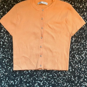 Orange ribbad topp från Zara - Kortärmad orange topp från Zara med ribbad struktur och söta blommiga knappar framtill. Modellen är croppad och har rund halsringning.  Använd en gång, mycket bra skick