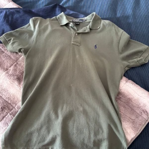 Olivgrön Polo t-shirt från Ralph Lauren - Snygg olivgrön t-shirt från Polo Ralph Lauren i klassisk pikémodell med krage och två knappar. Broderad logga i mörkblått på bröstet. Tillverkad i 100% bomull för skön känsla. Perfekt för dig som gillar stilrena och tidlösa plagg.
