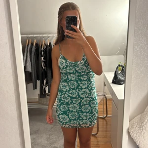 Grön blommig kortklänning från Monki - Superfin grön kortklänning från Monki med vita blommor, smala axelband och lätt draperad urringning. Sitter tajt och har en somrig vibe, perfekt för dig som vill sticka ut. Materialet känns svalt och mjukt mot huden.