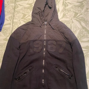 Svart hybridhoodie Polo Ralph Lauren, storlek M. - Svart hoodiejacka från Polo Ralph Lauren med quiltad ovandel och broderad 1967 på bröstet. Jackan har dragkedja framtill, två fickor med dragkedja och huva med snörning. Perfekt för dig som gillar streetwear och klassisk stil. Använd 2-3 gånger