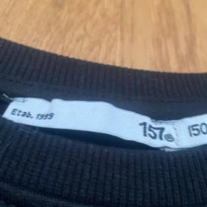 Svart sweatshirt från 157 - En klassisk svart sweatshirt från 157 med rund hals och ribbade muddar vid ärmslut och nederkant. Perfekt för dig som gillar enkel och stilren design. Tröjan är gjord i mjuk bomull och har långärmad passform som funkar året runt.