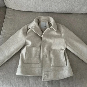 Beige kappa från lager 157 - Stilren beige jacka från 157 i ullmix med klassisk krage, stora framfickor och knäppning med tre knappar. Perfekt för dig som gillar en clean look och vill ha något som funkar till många outfits. Jackan har en rak passform och är helfodrad. Knappt använd, den är som ny!