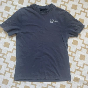 Mörkgrå t-shirt från Axel Arigato - Snygg mörkgrå t-shirt från Axel Arigato med vit tryckt text på bröstet. Klassisk rund hals och korta ärmar. Tillverkad i mjuk bomull för skön känsla. Perfekt för dig som gillar minimalistisk stil och streetwear vibes.