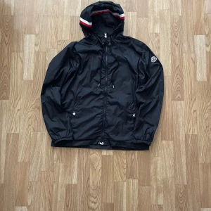 Svart windbreaker från Moncler  - Svart windbreaker från Moncler med huva som har röd och vit rand. Jackan har dragkedja framtill, två fickor med knappar och Moncler-logga på ärmen. Tillverkad i lätt och smidig polyester, perfekt för blåsiga dagar.