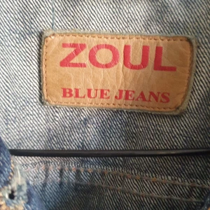 Mörkblå jeansjacka från Zoul - Snygg mörkblå jeansjacka från Zoul med klassiska knappar och kontrastsömmar. Jackan har två bröstfickor med lock och en röd broderad detalj på ena fickan. Tillverkad i 100% bomull för en autentisk denimkänsla.