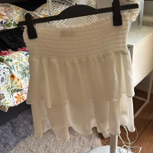 Vit volangkjol från Chelsea, stl M - Supersöt vit kortkjol från Chelsea i storlek M. Kjolen har bred smock i midjan och flera lager med volanger som ger en flowy och luftig känsla. Perfekt för dig som gillar romantisk och feminin stil. Materialet känns lätt och svalt, perfekt för varma dagar.