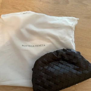 Brun axelväska från Bottega Veneta - Snygg brun axelväska från Bottega Veneta med klassiskt flätat mönster. Väskan är tillverkad i mjukt skinn och har en stilren, rundad form med smal axelrem. Perfekt för dig som gillar lyxiga detaljer och italiensk design.
