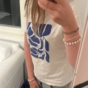 Vit t-shirt med blått grafiskt tryck - Snygg vit t-shirt med ett stort blått grafiskt tryck på bröstet. Modellen är klassisk med rund hals och korta ärmar. Perfekt till jeans eller shorts för en chill och modern look. Materialet känns mjukt och bekvämt mot huden.