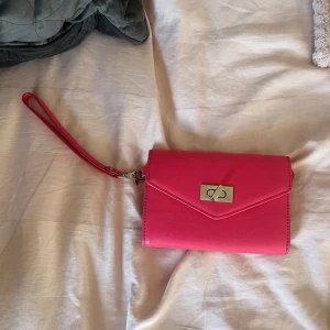 Rosa plånbok med handledsrem - Snygg rosa plånbok med avtagbar handledsrem och metallspänne framtill. Plånboken har flera fack för kort och ett fönster för ID eller foto. Stilren design med envelope-formad lock och struktur i materialet. Perfekt för att hålla koll på allt viktigt.