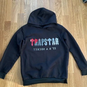 Svart Trapstar hoodie med tryck - Svart hoodie från Trapstar med stor logga i rött och grått framtill och texten 'IT'S A SECRET' under. Klassisk huva och ribbade muddar. Perfekt för dig som gillar streetwear och vill sticka ut med en fet grafisk detalj.