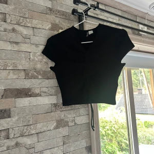 Svart ribbad croppad topp H&M Divided - Svart croppad topp från H&M Divided med ribbad struktur och v-ringning. Toppen har korta ärmar och snörning framtill som ger en snygg rynkad effekt. Perfekt att styla med höga jeans eller kjol för en trendig look.