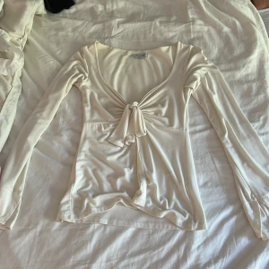 Vit omlottopp från Design by Si - Supersöt vit omlottopp från Design by Si med lång ärm och snygg knytning framtill. Toppens material är mjukt och stretchigt, perfekt för en trendig och feminin look. Djup v-ringning och lätt utsvängda ärmslut ger en extra touch. Obs lite knottrig på vissa ställen 