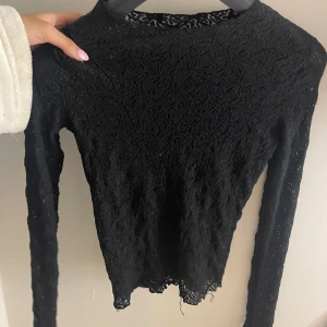 Svart spetsblus med lång ärm - Säljer en svart blus i spets med långärmad design och rund hals. Spetsmaterialet ger en snygg och lite romantisk vibe, perfekt att styla med jeans eller kjol. Blusen har en något vågig kant nedtill för extra detalj.