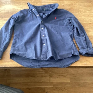 Blå skjorta från Ralph Lauren slim fit - Klassisk marinblå skjorta från Ralph Lauren i slim fit-modell. Skjortan har långa ärmar, knappar framtill och den ikoniska röda broderade loggan på bröstet. Tillverkad i mjuk bomull och passar perfekt till en stilren look.