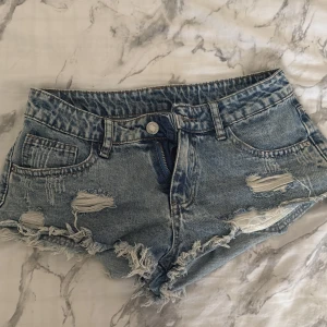 Blå slitna jeansshorts med fransar - Säljer ett par blå jeansshorts med slitna detaljer och fransig kant. Shortsen har klassisk femficksmodell, knapp och dragkedja framtill. Perfekta för sommardagar och ger en cool, avslappnad vibe. Köpta utomlands ❤️