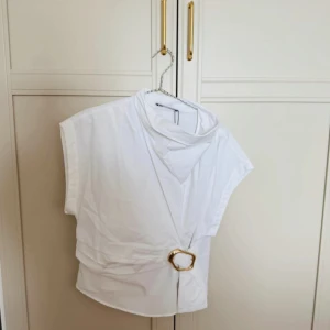 Vit omlottopp med gulddetalj från Zara - Snygg vit omlottopp från Zara med cool guldfärgad metallring i midjan. Toppen har korta ärmar och en hög krage som ger en trendig vibe. Perfekt att styla med jeans eller kjol för en chic look. Materialet är mjukt och lätt, vilket gör den skön att bära. Finns lite slitningar på emblemet 