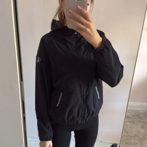 Svart vindjacka från Calvin Klein Jeans - Svart vindjacka från Calvin Klein Jeans med huva och dragkedja framtill. Jackan har logotyp på ärmen och vita tryck på fickorna. Modellen är loose fit med elastisk nederkant och ärmar. Materialet är lätt polyester med meshfoder för extra komfort.