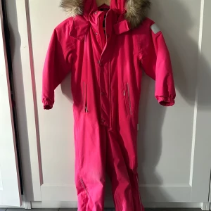 Rosa overall med fuskpäls på huvan - Snygg rosa overall från märket reima med huva som har fuskpäls, dragkedja framtill och praktiska fickor. Perfekt för kalla dagar med sin varma och vindtäta design. Elastiska muddar vid ärmarna och reflexdetaljer på axeln för extra synlighet. Storlek 110. Lite ljusare på knän & rumpa men fortfande hel. Använd en säsong