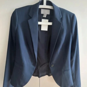 Mörkblå kavaj från H&M - Snygg mörkblå kavaj från H&M med klassisk krage och en knapp framtill. Figursydd modell med slät yta och diskreta fickor. Fodrad insida i matchande blått tyg. Perfekt för dig som gillar stilrena och enkla plagg