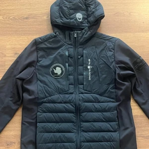 Svart pufferjacka från Sail Racing - Säljer en svart pufferjacka från Sail Racing med huva och dragkedja. Jackan har quiltade partier framtill och baktill samt logotyp på bröstet och ärmen. Perfekt för kyligare dagar och har en sportig look.