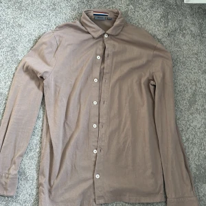 Beige skjorta från Snoot, storlek S - Stilren beige skjorta från Snoot i storlek S. Skjortan har klassisk krage, långa ärmar och vita knappar framtill. Tillverkad i mjukt bomullstyg som känns skönt mot huden. Perfekt för dig som gillar en enkel och clean look.