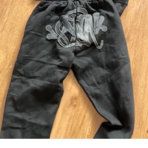 Svart hoodie och svart byxor från syna world - Svart hoodie från syna world med tryck på huvan och matchande svarta snören. Klassisk modell med lång ärm och luva, perfekt för en avslappnad stil.