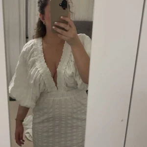 Vit kort omlottklänning med puffärm - Supersnygg vit kort klänning med djup v-ringning, puffiga trekvartsärmar och omlottdetalj. Klänningen har markerad midja och struktur i tyget som ger en lyxig känsla. Perfekt för sommarens alla tillfällen!