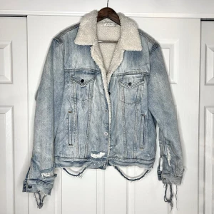 Ljusblå teddyfodrad jeansjacka MNML - Säljer en oversized ljusblå jeansjacka från MNML med vitt teddyfoder och krage. Jackan har slitna detaljer, knappar framtill och två bröstfickor. Perfekt för dig som gillar streetstyle och vill ha en jacka med attityd.