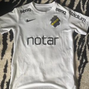 Vit AIK fotbollströja Nike. STORLEK 147-158 CM - AIK fotbollströja i vit polyester med svarta tryck och sponsorer som Notar, BenQ, Stadium, Fabege och SVEA. Klubbmärket sitter på bröstet och Nike-loggan är tryckt på framsidan. Nummer 12 på ryggen. Klassisk rund hals och korta ärmar.