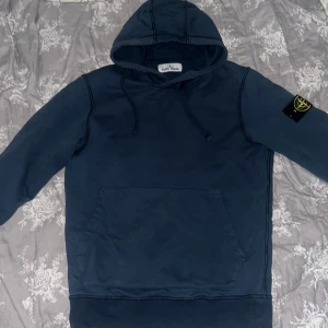 Mörkblå hoodie från Stone Island - Säljer en mörkblå hoodie från Stone Island med klassisk patch på ärmen och stor känguruficka framtill. Tröjan har huva med dragsko och är perfekt för en avslappnad stil. Märket syns tydligt i nacken och på etiketten.