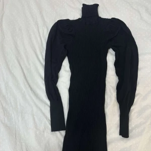Svart ribbad stickad klänning - Svart stickad klänning med ribbad struktur och höga puffärmar. Klänningen har polokrage och är figurnära med lång ärm. Perfekt för dig som gillar en stilren och trendig look.