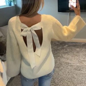 Krämvit stickad tröja med rosett - Superfin krämvit stickad tröja med öppen rygg och stor rosett bak. Tröjan har breda ärmar och en mjuk, ribbad struktur som känns både trendig och cozy. Perfekt för dig som vill ha något extra i vardagslooken.