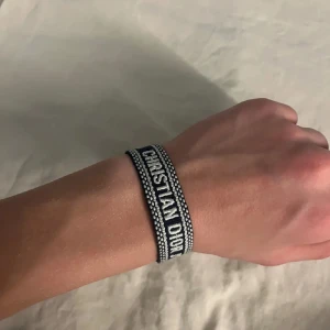 Christian Dior armband - Snyggt blått armband från Christian Dior med vit broderad text 'CHRISTIAN DIOR J'ADIOR'. Armbandet har ett coolt mönster med vita prickar och avslutas med två stiliga tofsar. Justerbar passform med knytning, perfekt för att ge din outfit en lyxig touch.