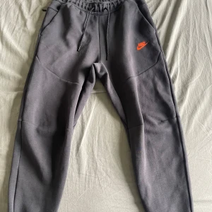 Mörkgrå nike tech byxor - mörkgråa nike tech byxor med orange logga på låret. lappen innanför byxan är avklippt. byxorna har använts ett par gånger men de är i bra skick. ursprungpris - 1100kr. 