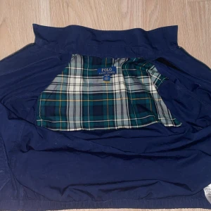 Blå vindjacka från Polo Ralph Lauren - Snygg marinblå vindjacka från Polo Ralph Lauren med dragkedja och klassisk vit logga på bröstet. Jackan har rutigt foder i grönt, blått och vitt, samt två fickor framtill. Perfekt för dig som gillar stilren och sportig look. Den är helt ny och har använt den 1-2 gånger, priset är inte helt hugget i sten, NYPRIS 2800!