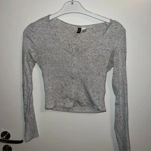 Grå croppad långärmad topp H&M - Gråmelerad croppad topp från H&M med lång ärm och v-ringning. Toppen har knappar framtill och är i en mjuk, tunn bomullsblandning. Perfekt till jeans eller kjol för en chill och enkel look.