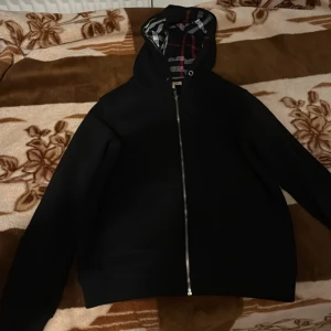 Svart Burberry hoodie med dragkedja - Svart hoodie från Burberry med klassiskt rutigt foder i huvan och insidan, dragkedja framtill och långa ärmar. Snygg och stilren design med ikoniska Burberry-rutor i svart, vitt och rött. Perfekt för dig som vill ha en exklusiv streetwear-look.