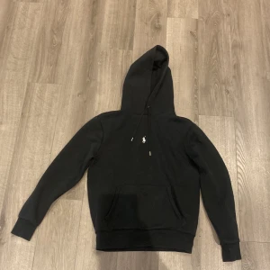 Svart hoodie från Ralph Lauren - Svart hoodie från Ralph Lauren med klassisk vit broderad logga på bröstet. Tröjan har huva med snörning, känguruficka framtill och ribbade muddar vid ärmslut och nederkant. Perfekt för en chill och stilren look.