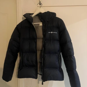 Svart dunjacka från Sail Racing - Snygg blå dunjacka från Sail Racing med huva och dragkedja. Insidan är ljusgrå och jackan är perfekt för kalla dagar. Passformen är normal och jackan har långa ärmar. Lite smink på insidan som inte märks när jackan är på. Priset kan diskuteras!!