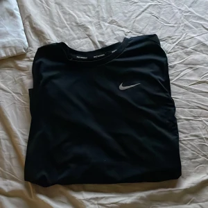 Svart Nike Dri-Fit långärmad tröja - Nike Dri-Fit långärmad tröja i svart med diskret logga på bröstet. Perfekt för löpning eller träning, tillverkad i lätt och ventilerande polyester som håller dig torr. Snygg och enkel design med rund hals och normal passform. Storlek L men passar M