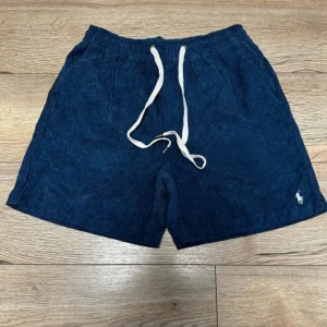 Blå manchester shorts från Polo Ralph Lauren - Snygga mörkblå manchester shorts från Polo Ralph Lauren med vit broderad logga på benet. Shortsen har elastisk midja med vit snörning och sidfickor. Perfekta för en chill sommarlook med skön loose passform och klassisk stil. Storleken passar på M (W30-W32). RL taggarna är fortfarande kvar på kläderna.  