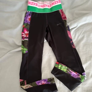 Nike Pro Dri-FIT leggings med blommor - Nike Pro Dri-FIT leggings i svart med bred grön och rosa resår i midjan och färgglada blommiga paneler längs sidorna och bensluten. Sitter tight och är perfekta för träning. Materialet är polyester som andas och transporterar bort fukt.