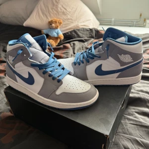 Nike Jordan 1 Mids, true blue - Nike Air Jordan 1 Mid (true blue) sneakers i fräsch vit, blå och grå färg. Klassisk high-top siluett med blå snörning, rund tå och platt sula. Swoosh-logga i blått på sidan och Air Jordan Wings på ankeln. Ovandel i skinn och syntet, insida i blå mesh för extra komfort. Storlek 45, endast används ett fåtal gånger! Box ingår!