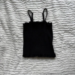 Svart smockad linnetopp från XOXO Friends - Svart smockad linnetopp från XOXO Friends med tunna axelband och volangkant nertill. Toppen är gjord i mjuk bomull med lite stretch för en bekväm passform. Perfekt att matcha med jeans eller kjol för en cool look.