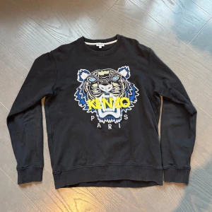 Svart Kenzo sweatshirt med tigertryck - Svart sweatshirt från Kenzo med ikoniskt broderat tigerhuvud i blått, vitt och gult framtill. Tröjan har rund hals, ribbade muddar och är långärmad. Perfekt statement-plagg för dig som vill sticka ut med en cool streetstyle-look. Några Småhål men inget som syns vid användning 